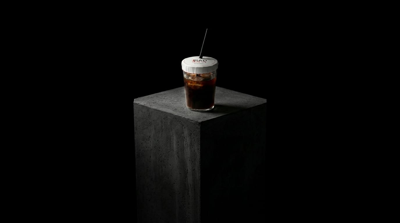 Vaso con hielo y refresco cerrado por arriba con la tapa adhesiva BadIdea, sobre un pedestal de hormigón con fondo negro
